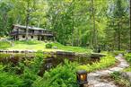Modern Rustic Retreat with Wraparound Views & Water FeaturesWelcome to this exceptional modern-rustic home that seamlessly blends mountain-lodge charm with updated comfort and style. Set back from the road, a long private driveway lined by an extensive, hand-built rock wall leads you to this striking residence â€” offering both curb appeal and a sense of arrival.Step onto the dramatic wraparound deck, where youâ€™ll enjoy tranquil views of a picturesque pond, complete with several charming footbridges crossing a scenic, running brook that winds through the property. Itâ€™s a rare outdoor setting that offers peace, beauty, and a strong connection to nature.Inside, the home features expansive windows, and newly finished red oak floors that fill the interior with warmth and natural light. 2 stone fireplaces anchoring the open-concept living area, creating a cozy yet spacious atmosphere.The lower level provides flexible living space, ideal for a guest suite, media room, or home office, with direct access to the two-car garage and the surrounding grounds.Recent Updates & Features:â€¢ Hand crafter wood box lighting throughout whole propertyâ€¢ Brand-new air conditioning systemâ€¢ Modern water treatment systemâ€¢ New water heater (2023)â€¢ Newly sanded and refinished red oak floorsâ€¢ Builder owned, crafted and architectedâ€¢ 2 sheds â€“ attached and a large detached â€¢ 2 car garageâ€¢ Wraparound deck overlooks pond and brookâ€¢ Several footbridges cross the scenic running broâ€¢ Meticulously landscaped, breathtaking groundsâ€¢ Mid-century modern and rustic architectureâ€¢ Serene wooded setting just minutes from town, lakes, or trail systems